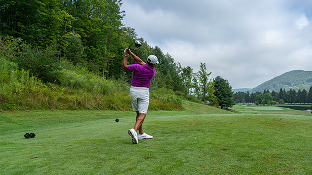 A man tees off at the Double Black Diamond Golf Course with scenic hills in the background.